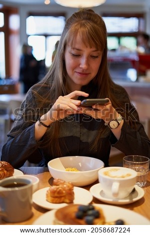 Beautiful young female student taking photos of sweet dessert and flat white coffee on smart phone on cafe. Trendy millennial caucasian blogger having breakfast in modern coffeeshop