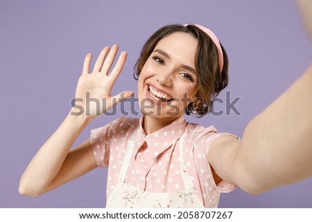 Close up friendly blogger happy young housewife housekeeper chef cook baker woman wear pink apron do selfie shot on mobile phone waving hand isolated on pastel violet background. Cooking food concept