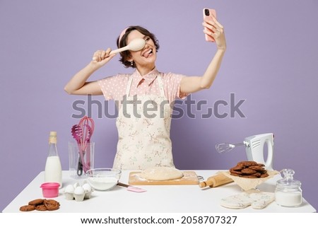 Young fun housewife chef baker woman in pink apron work at table kitchenware do selfie shot mobile cell phone cover eye with spoon isolated on pastel violet background Process cooking food concept.