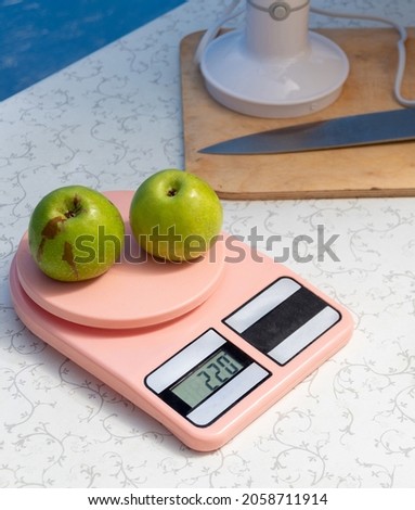 Two green apples are lying on the kitchen scales. There is a knife and a blender on the table nearby. Selective focus.