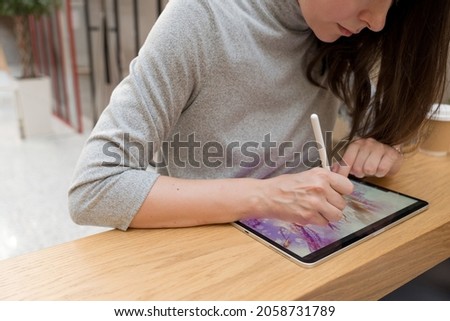 A digital artist draws on a graphic screen tablet using a stylus, a freelancer works in a public space coworking, the ability to work remotely. A young woman artist or designer uses a tablet to draw Royalty-Free Stock Photo #2058731789