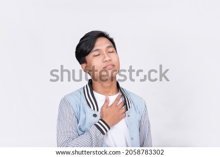 A young Filipino breaths a sigh of relief. Hands on chest and eyes closed. Isolated on white background. Royalty-Free Stock Photo #2058783302