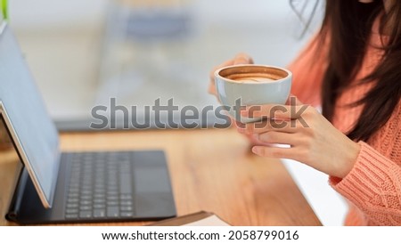 Cropped closeup image of Young female in pink sweater drinking, holding hot coffee cup while working on tablet at the cafe