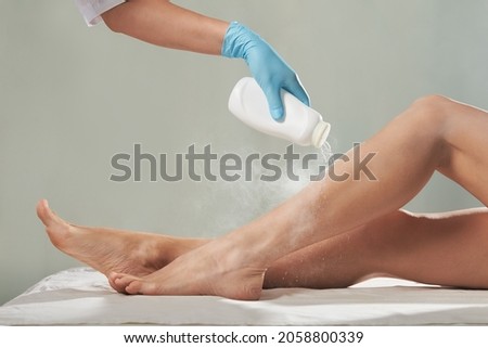 Cosmetologist is sprinkling and distributing talcum powder on a young girl's leg before the epilation procedure. The girl is lying on a couch in a beauty salon, she does the procedure shugaring Royalty-Free Stock Photo #2058800339