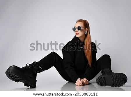 Cool trendy rich red-haired woman rock star, biker in modern black hoodie, pants and massive brutal army shoes sits on floor with legs wide apart and looks aside at copy space Royalty-Free Stock Photo #2058803111
