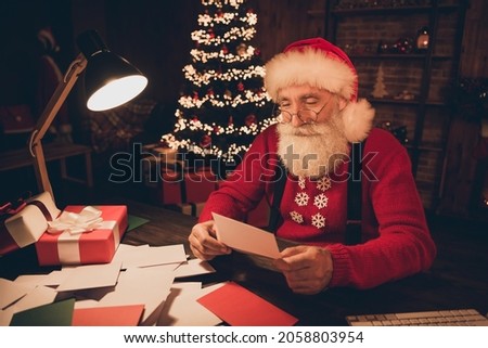Photo of cheerful nice santa claus sit table dark evening read letter wear hat sweater in north pole office indoors