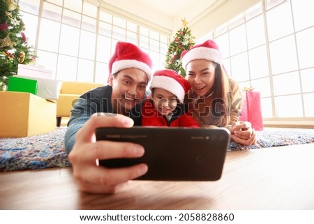 Asian Selfie holding smartphone Taking picture. Happy young family with kids fun celebrating Christmas. Christmas time. My dad, mom, and daughter in Santa hats lie about In front Gift box at home