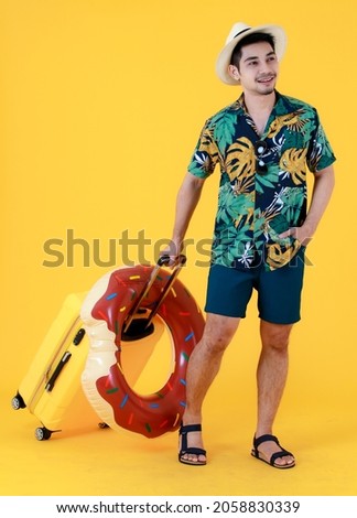 Happy young Asian man wearing colorful printed Hawaiian shirt and panama hat pulls yellow suitcase and swim ring. Full body studio portrait on yellow background. Summer holiday travel concept