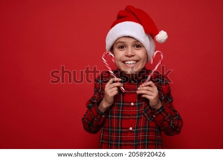 Christmas and New Year celebration concept with space for ad. Funny amazing handsome child boy in Santa hat rejoices looking at camera, holding sugary striped candy canes, sweet lollipops in his hands