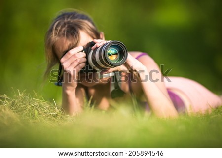 Pretty female photographer lying in grass, on a lovely summer day, taking pictures with her DSLR camera and a telephoto lens