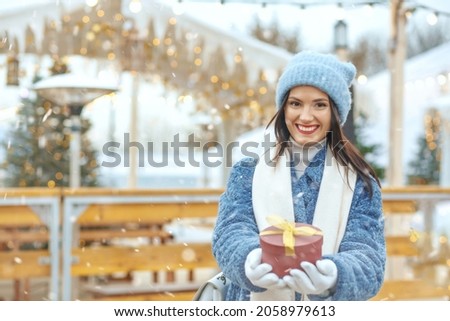 Joyful brunette woman in winter coat holding a gift box at christmas fair during the snowfall. Space for text