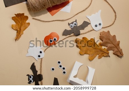 homemade garland on a jute rope made of pumpkins, bats and ghosts with eyes on a beige background next to scraps of colored paper, leaves and eyes Royalty-Free Stock Photo #2058999728