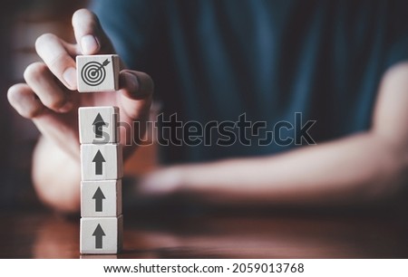 Businessman holding wooden cube with target board icon and arrow on wooden table. Goals and planning for success in marketing business, achieve the objective concept. Closeup and free copy space