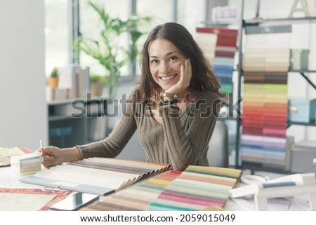 Young interior designer sitting at desk and working in her studio, she is smiling at camera