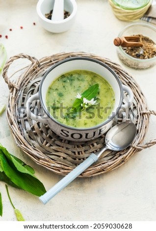 Wild garlic soup in white bowl on wooden plate with spoon and wild garlic leaves at kitchen table with bowl and mortar and pestle. Cooking healthy lunch at home with seasonal herbs. Table top view. Royalty-Free Stock Photo #2059030628