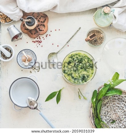 Wild garlic soup on white kitchen table with bowls, ingredients, herbs, spices and kitchen utensils. Cooking with seasonal wild herbs at home. Top view.