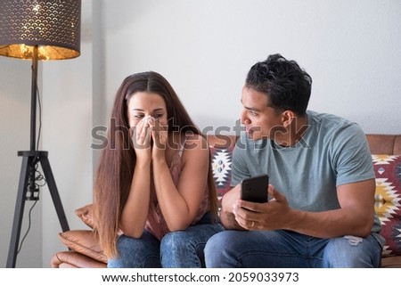 Boyfriend asking for explanation about phone cellular messages tf her girlfriend sitting on a couch in the living room in a house interior with a dark background - lie and couple problems concept Royalty-Free Stock Photo #2059033973
