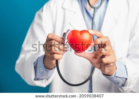 Hand of a doctor holding a stethoscope checking a red heart shape while standing in the hospital or clinic. Close-up photo. Space for text. Medical, treatment, and healthcare concept.
