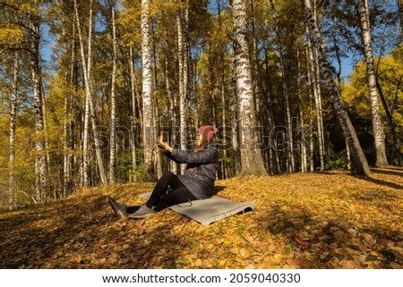 Young beautiful woman sits on a rug in the forest and holds a mobile phone in her hands. The girl in the autumn forest sits on the ground. A woman in a birch grove takes a selfie.