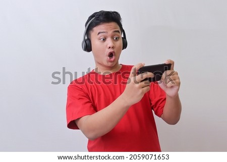 Portrait of attractive Asian man in red t-shirt using wireless headphone playing games on his mobile phone by tilting the screen. Wow face expression. Isolated image on gray background Royalty-Free Stock Photo #2059071653