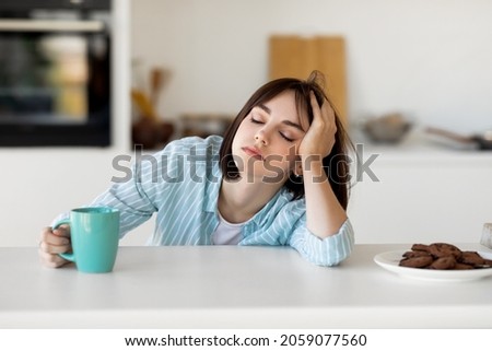 Sleepy young woman drinking coffee, feeling tired, suffering from insomnia and sleeping disorder. Sad female sitting in modern kitchen interior, empty space Royalty-Free Stock Photo #2059077560