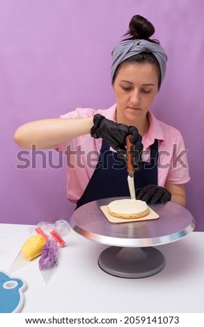 A woman in an apron and gloves on her hands, applies cream to the first cake of a bento cake with a scraper. The assembly process. Selective focus.
