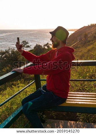 Bearded millennial man in red hoody backpack on bench with smartphone taking photo of autumn sea view. Authentic male tourist Blogger guy lifestyle Solo travel adventure. Blogging walking backpacking.