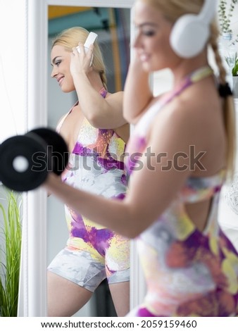 Enjoying fitness. Sportive woman. Home training. Perfect body. Happy athletic lady in colorful sportswear enjoying dumbbells workout listening music headphones mirror reflection light room interior.