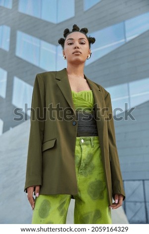 Vertical shot of thoughtful self assured young woman with trendy hairstyle bright makeup dressed in fashionable apparel poses against grey wall outside. Youth urban lifestyle and fashion concept Royalty-Free Stock Photo #2059164329