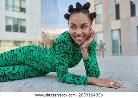 Horizontal shot of positive teenage girl with combed hair buns bright makeup smiles toothily looks away dressed in green costume poses outdoors against modern city building poses for making photo
