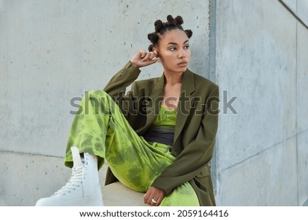 Horizontal shot of beautiful teenage girl with trendy hairstyle dressed in green clothes looks away poses against urban grey wall considers something. People youth lifestyle and fashion concept Royalty-Free Stock Photo #2059164416