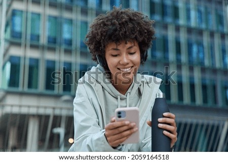 Positive sporty woman drinks water after exercising uses mobile phone scrolls social networks smiles happily dressed in hoodie and anorak poses against modern city building enjoys messaging. Royalty-Free Stock Photo #2059164518