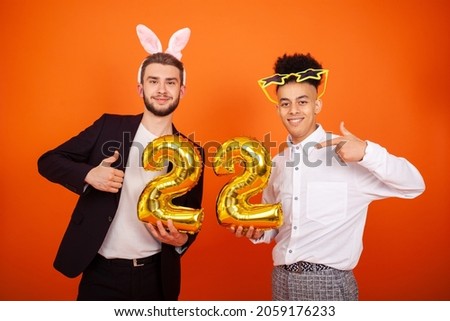 Celebration, party and people concept - Two funny international friends in bunny ears and star glasses, showing thumbs up and holding a number 22 shaped balloon on an orange background with copy space