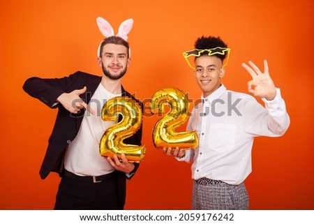 Celebration, anniversary and people concept - Two joyful young friends wearing bunny ears and big star glasses, celebrating a birthday and holding a number 22 shaped balloon on an orange background
