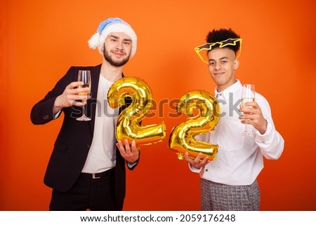 Celebration, party, anniversary and people concept - Happy Caucasian and American in a blue Santa hat and star glasses holding a number 22 shaped balloon and champagne glasses on an orange background