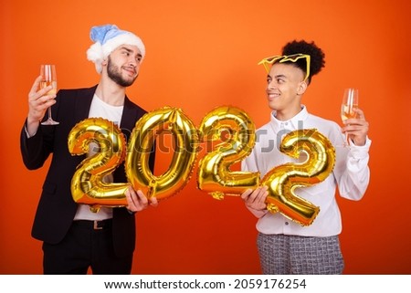 New year, lgbt, celebration and people concept - Happy international gay men looking at each other with star glasses and Santa hat, holding a number 2022 and champagne glasses on an orange background