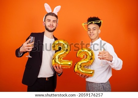 Celebration, party, anniversary and people concept - Two cheerful international friends in bunny ears and star glasses holding a number 22 shaped balloon and champagne glasses on an orange background