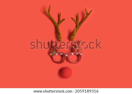 Funny Christmas face with antlers of a deer, toy glasses and a clown nose on a red background. Christmas or New Year party concept. Flat lay. Top view