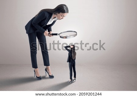 Businesswoman in suit using magnifier magnifying glass to look at tiny subordinate on concrete background with shadow and mock up place. Hiring and dominance concept