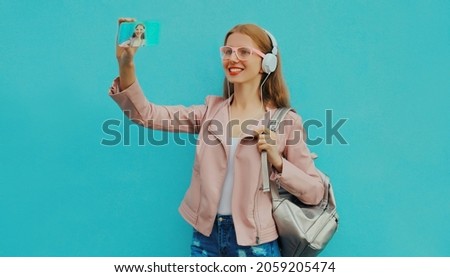 Portrait of modern young woman taking selfie by smartphone in headphones listening to music on blue background