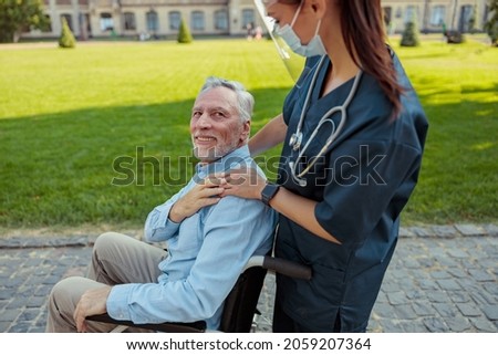 Caring nurse wearing face shield and mask on a walk with aged man, recovering patient in a wheelchair on a summer day outdoors