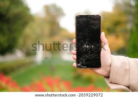 Broken glass on the phone screen. Broken phone in a woman's hand. Background from plants in the park. Blurred background