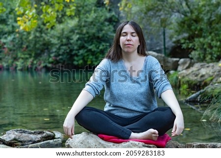 young brunette girl doing yoga and meditation on a high mountain river with a lake in the background  wearing blue sweater and black pants