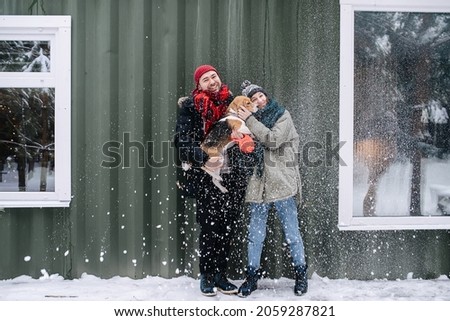 Cute couple posing for a photo with their beagle outdoors at winter. Man is holding the dog in his arms, woman is leaning to her. In front of corrugated sheet house.