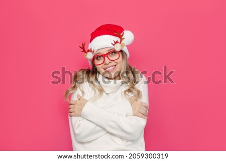 A young frozen charming smiling blonde woman in glasses with deer antlers, a white warm sweater and Santa hat enjoys the softness of the material hugging herself isolated on a color pink background