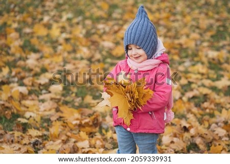 Sweet little girl enjoys autumn. The child plays in the autumn forest and throws leaves. A girl in a pink jacket and a gray hat against the background of autumn leaves