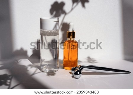 Bottle with clear micellar water, massager and oil on a white background with a shadow from flowers. Lotion for cleansing the skin and eyes from cosmetics, makeup. Beauty product in plastic packaging