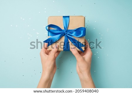 First person top view photo of hands holding craft paper giftbox with vivid blue ribbon bow over shiny sequins on isolated pastel blue background Royalty-Free Stock Photo #2059408040