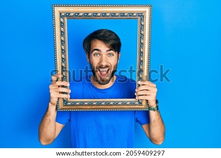 Young hispanic man holding empty frame smiling and laughing hard out loud because funny crazy joke. 