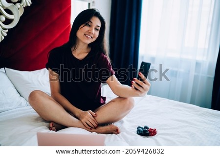 Smiling Asian hipster girl making selfie pictures during leisure weekend in home bedroom, happy female blogger shooting video vlog via cellphone app using wifi internet connection in hotel